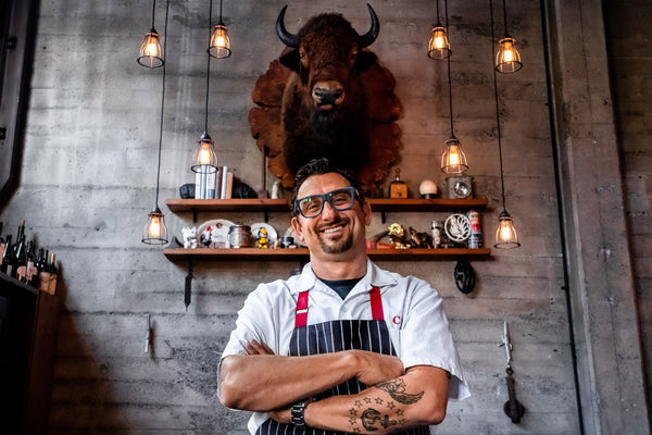 Chris posing, arms folded with a broad smile, in front of a mounted cow's head on a concrete interior wall, lit with several low-wattage hanging lightbulbs.