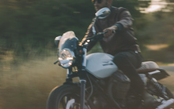 Blurred shot of a man riding a motorcycle, wearing a brown leather jacket.